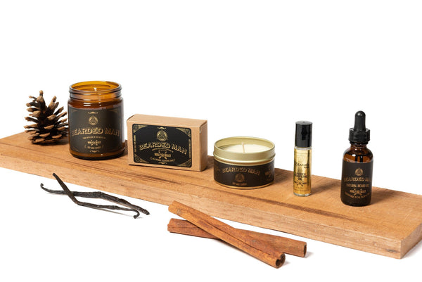 Bearded Man fragrance collection of soy candles natural bar soap beard oil perfume oil sitting on a cedar board with pine cone, vanilla bean, and cinnamon sticks