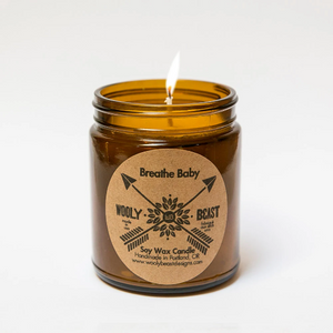 lit cotton wick soy candle burning in glass amber apothecary jar with circle kraft paper label on white background. label reads Breathe Baby wooly beast crossed arrow logo soy wax candle handmade in portland oregon