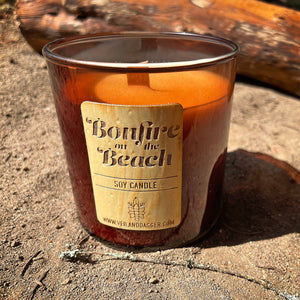 Bonfire on the beach scented soy candle by Veil and Dagger Fine Home Goods sitting on sand by driftwood on the beach