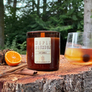 Maple Bourbon Soy Candle in amber tumbler jar with wood wick on tree stump in forest with slice orange and Old Fashioned cocktail pine cone cinnamon sticks by Veil and Dagger fine home goods