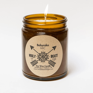 babycake soy candle with cotton wick burning in amber apothecary jar on white background circle kraft paper label design has crossed arrows and reads Babycake NYC wooly beast soy wax candle handmade in portland,  or