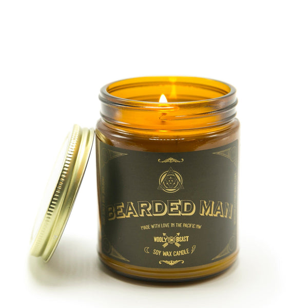 Bearded man scented soy candle burning in amber glass apothecary jar with gold lid on white background black and gold label design has sacred geometry and says bearded man made with love in the pacific nw wooly beast soy wax candle