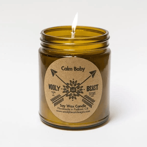 candle burning in amber glass apothecary jar with cotton wick on white background. Circle kraft paper label has crossed arrows and reads Calm Baby wooly beast soy wax candle handmade in portland oregon