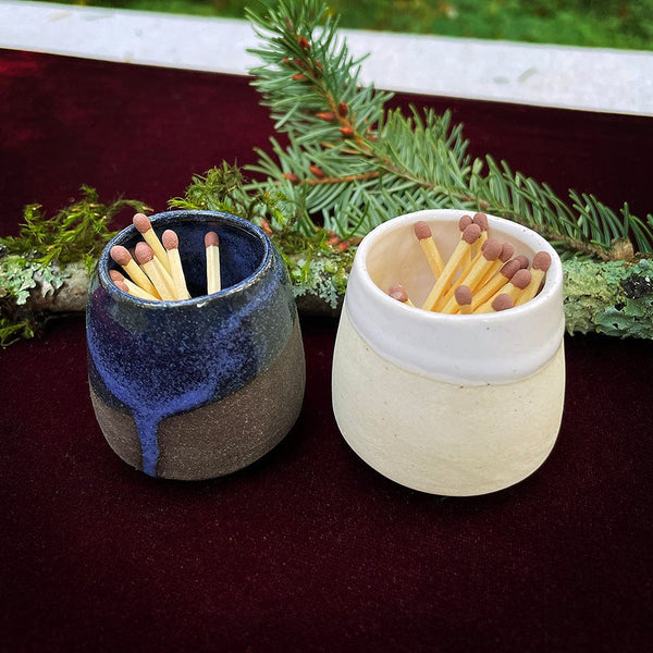 handmade ceramic toothpick matchstick holders in blue dark grey and white cream colors holding red tipped matchsticks on a red velvet fabric with douglas fir branch on tabletop