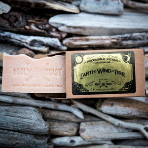 handmade essential oil soap bar coming out of kraft paper box package, lying on driftwood, pink soap bar has wooly beast crossed arrow logo soap stamp, black and gold label has oujia board design and reads supernatural mystical cleansing bar earth wind and fire wooly beast all natural mystical soap