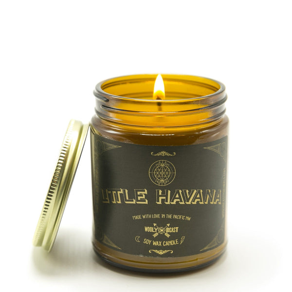 soy candle burning in amber glass jar with gold lid leaning against it on white background. Black and gold label has sacred geometry design and reads Little Havana soy wax candle made with love in the pacific nw wooly beast logo with crossed arrows