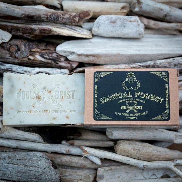 magical forest natural white bar soap coming out of kraft paper box packaging, lying on top of driftwood. Black and gold label has triple goddess symbol and reads Magical Forest handmade with love in portland, or wooly beast crossed arrow logo, all natural cleansing soap