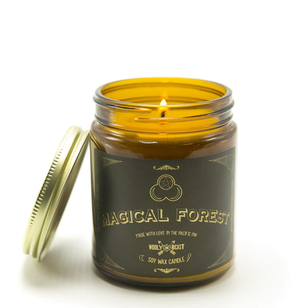 Magical Forest scented Soy Candle burning with gold lid leaning against the amber glass jar on white background, label design has triple moon goddess symbol