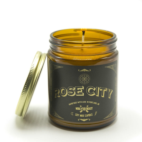 Rose City soy candle wick burning with gold lid leaning against side on white background. Black and gold label has sacred geometry design and says rose city handmade with love in portland, oregon wooly beast soy wax candle