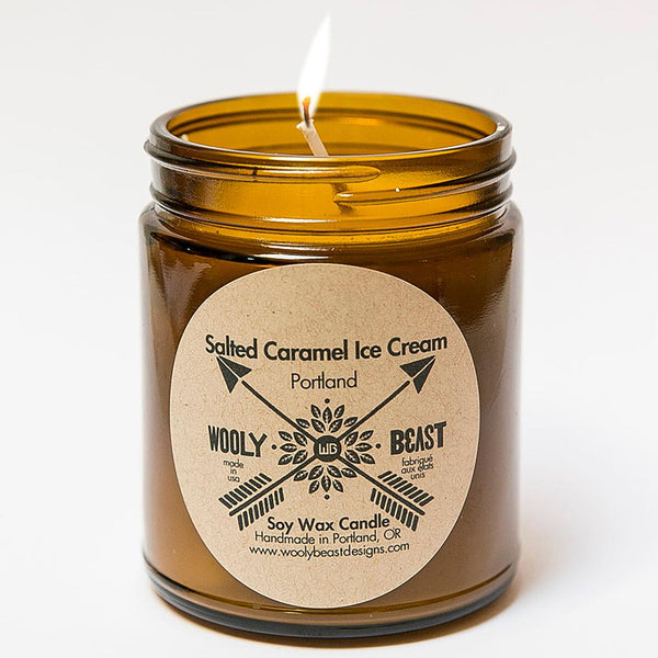 salted caramel ice cream soy candle in amber jar, cotton wick burning, on white background. circle kraft paper label reads salted caramel ice cream portland wooly beast crossed arrows logo, soy wax candle, handmade in portland oregon