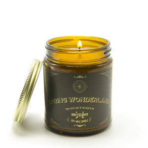 lilac and hydrangea soy candle burning in amber glass jar with gold lid leaning against it on white background. Black and gold label has sacred geometry design and reads Spring Wonderland soy wax candle made with love in the pacific nw