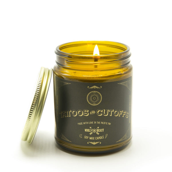Tattoos and Cutoffs Soy Candle wick burning in amber glass apothecary jar with gold lid leaning against it, black and gold label has sacred geometry design and reads Tattoos and Cutoffs made with love in the pacific nw wooly beast soy wax candle