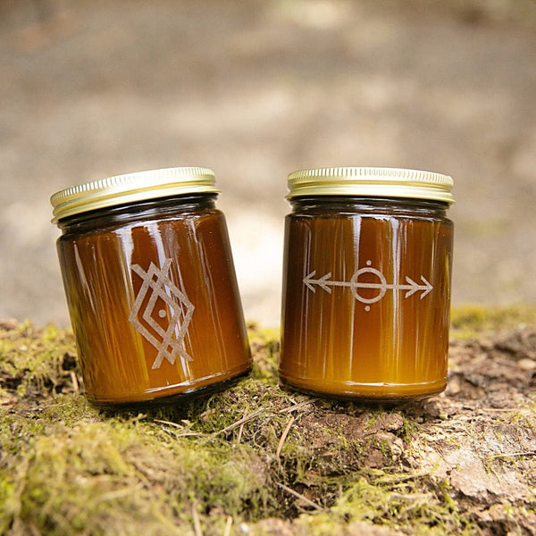 the woodsman scented soy candle laser engraved nordic runes design in amber glass jars with gold lids sitting on mossy log