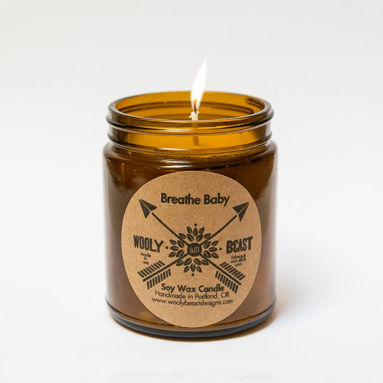 lit cotton wick soy candle burning in glass amber apothecary jar with circle kraft paper label on white background. label reads Breathe Baby wooly beast crossed arrow logo soy wax candle handmade in portland oregon