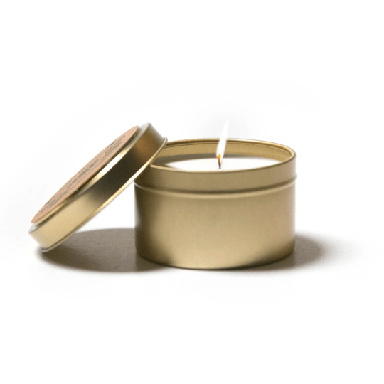 lit cotton wick soy candle burning in gold metal tin container with gold metal lid leaning against it on white background
