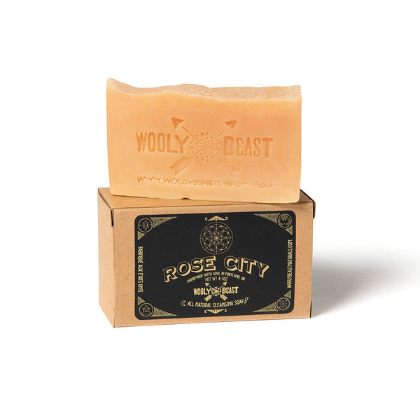 pink colored Rose City fresh cut roses natural bar soap  on top of kraft paper box on white background. Black and gold label has sacred geometry design and reads Rose City handmade with love in portland oregon Wooly Beast crossed arrows logo, all natural cleansing soap