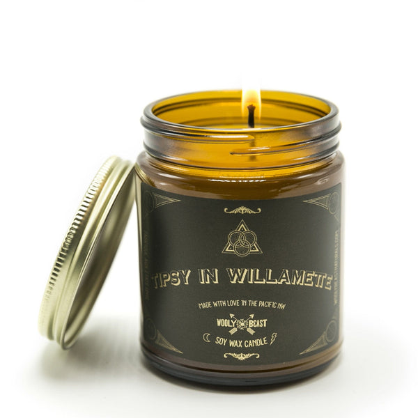 tipsy in willamette soy candle lit wick burning in amber apothecary jar with gold lid leaning against it on white background black and gold label has sacred geometry design and reads tipsy in willamette made with love in the pacific nw wooly beast soy wax candle