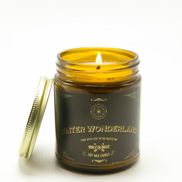 winter wonderland soy candle wick burning with gold lid leaning against amber glass apothecary jar on white background. black and gold label has sacred geometry design and reads winter wonderland made with love in the pacific nw wooly beast soy wax candle